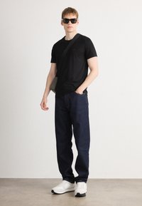 Black crew-neck t-shirt, dark blue jeans, and white sneakers. Model wears black sunglasses and carries a gray crossbody bag.