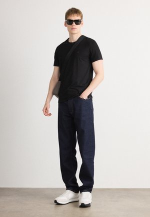 Zwarte crew-neck t-shirt, donkerblauwe spijkerbroek en witte sneakers. Model draagt een zwarte zonnebril en heeft een grijze crossbody tas.