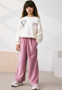 Biely sweatshirt s grafikou kresleného charakteru a dúhovým detailom, v kombinácii s voľnými ružovými velúrovými nohavicami a šedými teniskami.