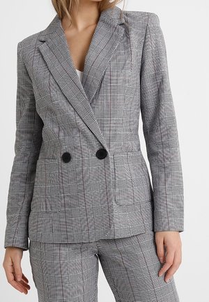 Mujer que lleva un blazer cruzado de cuadros gris ajustado y pantalones a juego con botones negros en la parte delantera del blazer.