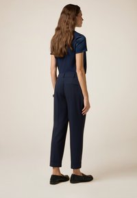 Donna con lunghi capelli castani ondulati, che indossa una maglietta a maniche corte blu navy, pantaloni tagliati blu navy fino alla caviglia e mocassini neri, in piedi su uno sfondo beige.