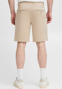 Shorts beige avec une texture lisse, deux poches arrière avec fermetures à bouton, et une coupe droite, associés à des chaussettes blanches et des baskets.