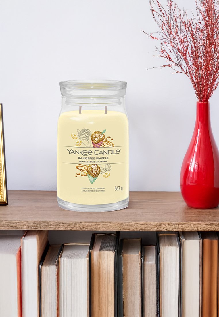 Świeca zapachowa Yankee Candle Banoffee Waffle w słoiku na drewnianej półce, z książkami poniżej oraz czerwoną wazą z suchymi gałązkami obok.