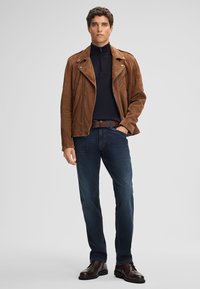 Bruine suède motorjack met een ritssluiting aan de voorkant, zwarte coltrui, blauwe slim-fit jeans en bruine leren schoenen, staand tegen een grijze achtergrond.