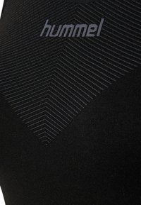 Fekete sportfelső, amely texturált geometrikus mintával és szürke "hummel" logóval rendelkezik. Simább anyag, testhezálló fazon és minimalista stílus.