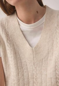 Beige Strickweste mit V-Ausschnitt und Kabelmuster. Getragen über einem weißen kurzärmeligen Hemd mit gerippter Textur am Hals.