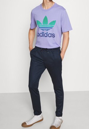 Mann trägt ein hellviolettes Adidas-T-Shirt, dunkle Hose mit Kordelzug und weiße Sneakers, steht mit den Händen in den Taschen.