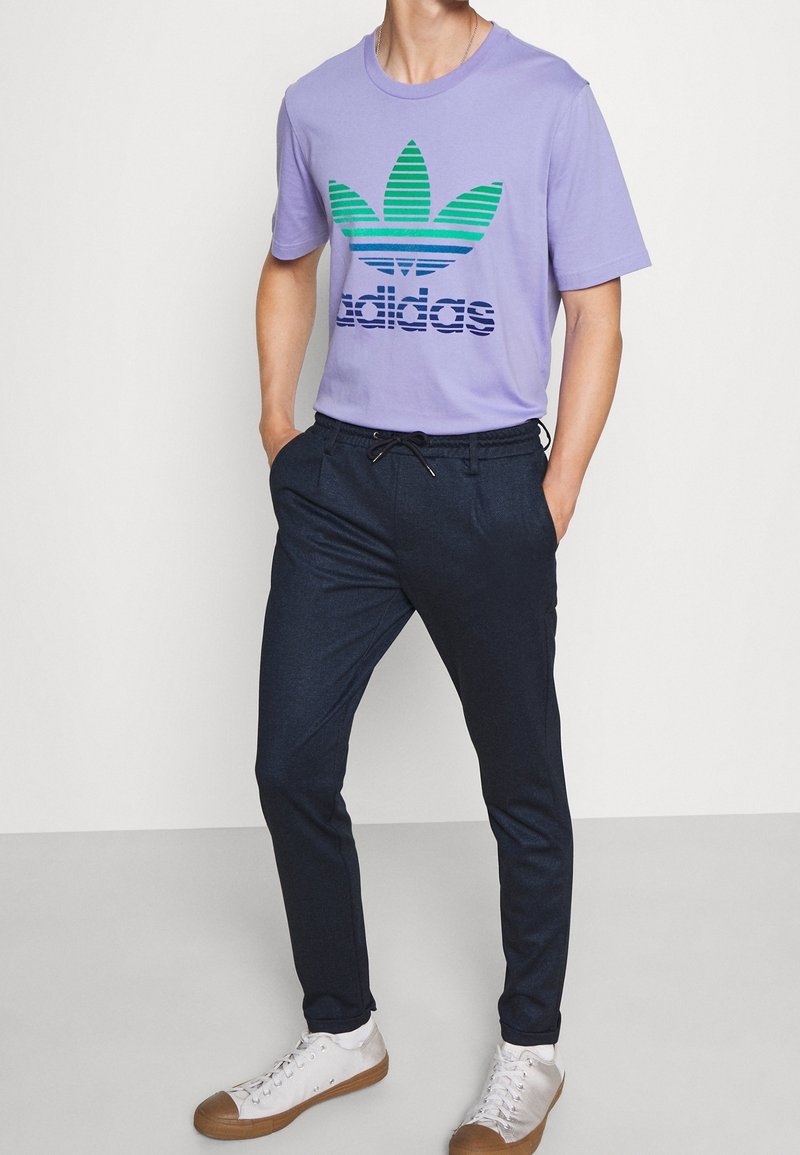 Homme portant un t-shirt Adidas violet clair, un pantalon sombre avec cordon et des baskets blanches, debout les mains dans les poches.