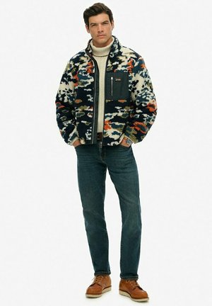 Fleece jacket with a multicolored camouflage pattern in blue, orange, and white; features a zip front and pocket. Paired with blue jeans and brown shoes.