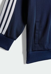 Mornarska športna jakna z rebrastimi manšetami in robom, ki ima tri bele črte vzdolž ramena in en stranski žep. Gladka tekstura.