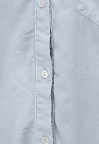 Jasnoszara, tkana koszula z dekoltem zapinanym na guziki, pionowymi szwami i białymi plastikowymi guzikami na listwie. Gładka tekstura.