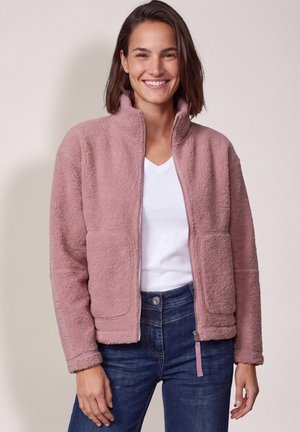 Femme souriante portant une veste polaire rose zippée par-dessus une chemise blanche et un jean bleu sur un fond clair uni.