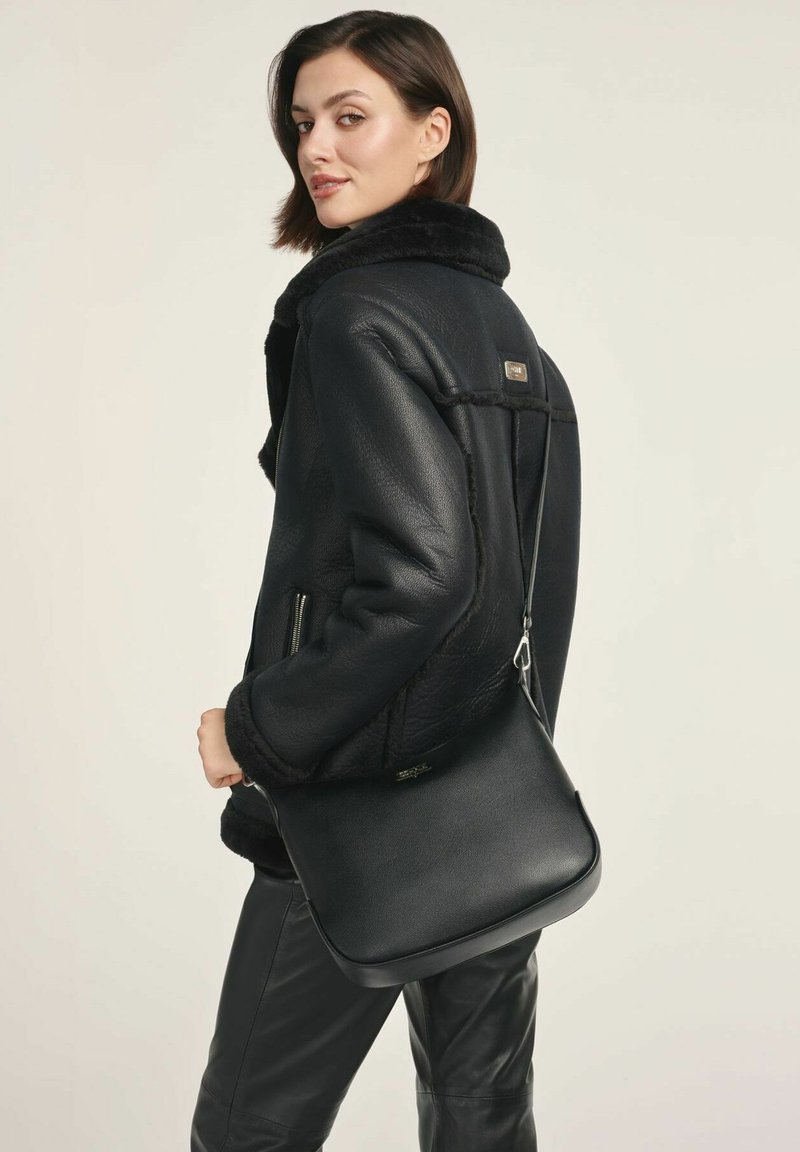 Kobieta w czarnej skórzanej kurtce shearling i czarnych skórzanych spodniach trzyma czarną skórzaną torebkę na pasku, patrząc przez ramię.