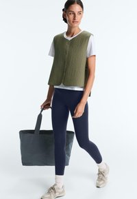 Frau trägt eine olivgrüne gesteppte Weste über einem weißen T-Shirt und marineblaue Leggings, trägt eine große dunkelgraue Tragetasche.