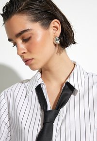 Randig vit skjorta med svart slips, med en slät textur. Stora silverörhängen med ett marmormönster. Håret slickat bakåt.