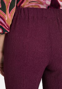Pantalons bordeaux texturés, avec une taille élastique, présentant des détails en côtes verticales. Le tissu semble doux et extensible.