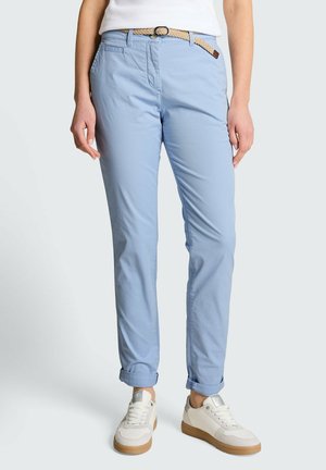 Kobieta ubrana w jasnoniebieskie podwinięte spodnie z beżowym plecionym paskiem i białe trampki z gumowymi podeszwami, stojąca na jednolitym tle.