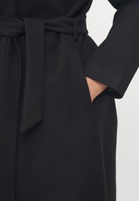 Cappotto nero realizzato in tessuto testurizzato, dotato di una cintura legata in vita e di una tasca laterale con dettagli di manica a polsino.