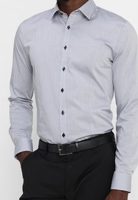 Vaaleanharmaa napitettava paita, jossa on jämäkkä kaulus, mustat napit ja hienovarainen tekstuuri, yhdistettynä mustiin housuihin ja vyöhön.