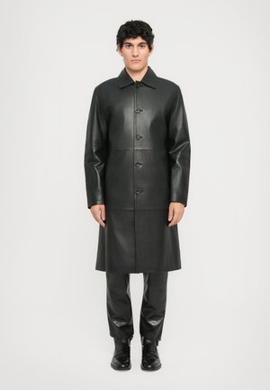 Man standing wearing a long black leather buttoned coat, black textured pants, and black leather shoes against a plain white background.
