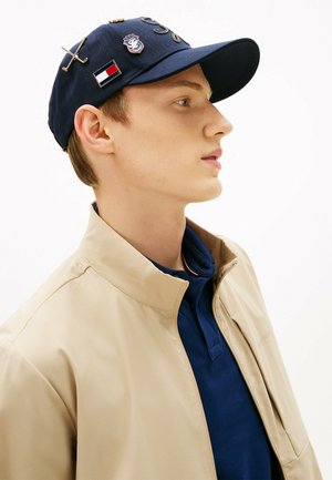 Jonge man in beige jas en donkerblauw poloshirt draagt een donkerblauwe pet met golf-gerelateerde spelden op een witte achtergrond, kijkt naar rechts in profiel.