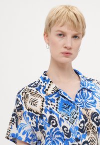 Camisa de colarinho de mangas curtas com um padrão floral azul, preto e bege, feita de tecido leve com uma textura suave.