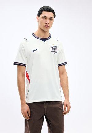 Jeune homme portant un maillot de football d'Angleterre avec le logo Nike et le blason des trois lions, debout devant un fond blanc uni.