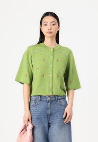 Grønn strikket cardigan med korte ermer, med blomsterbroderi i rosa og blått, og fem hvite knapper foran.