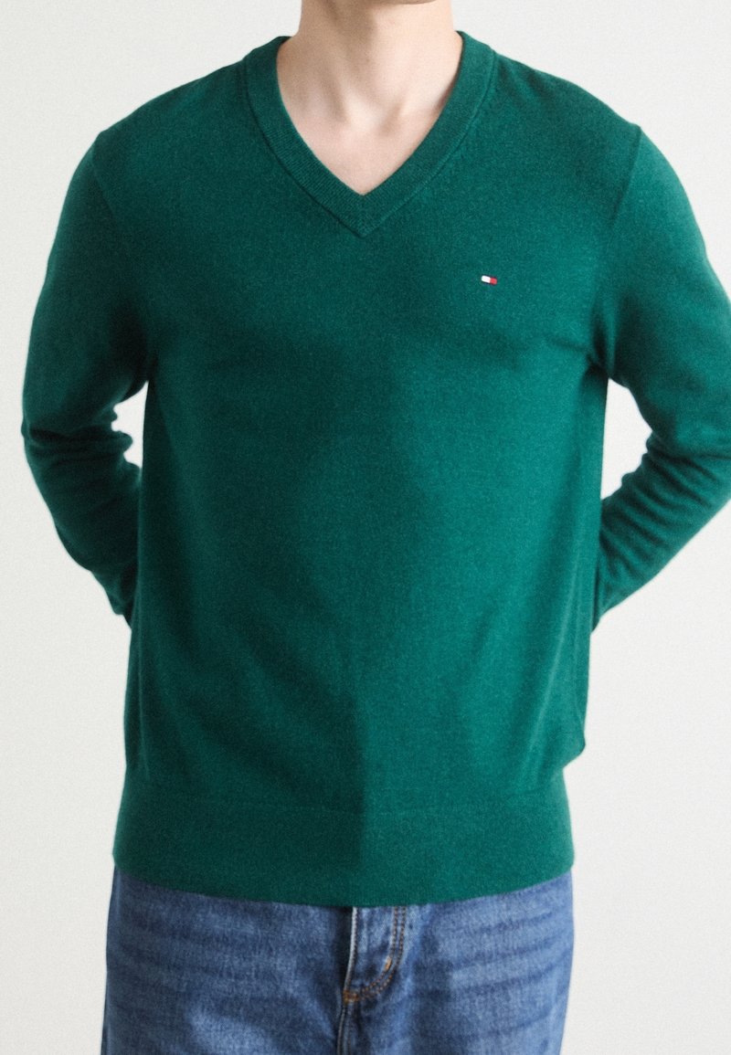 Personne portant un pull vert à col en V avec un petit logo rouge, blanc et bleu sur la poitrine et un jean bleu, debout les mains derrière le dos.
