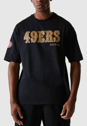 Mann trägt ein schwarzes, übergroßes T-Shirt mit großem goldenen Aufdruck "49ERS EST. 1946" auf der Vorderseite und einem roten Logo-Patch auf dem linken Ärmel.