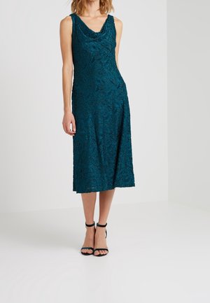 Femme portant une robe midi sans manches couleur sarcelle avec un motif floral texturé, associée à des escarpins noirs à bride cheville, debout devant un fond blanc.