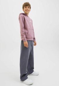 Ljust rosa hoodie med känguruficka och logotyp, kombinerad med löst sittande mörkgrå byxor och vita sneakers med låg profil.