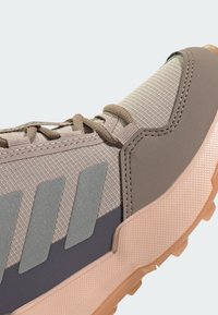 Beige athletic shoe featuring a textured upper, dark gray accents, and three reflective stripes. Rubber sole with a patterned tread for grip.
