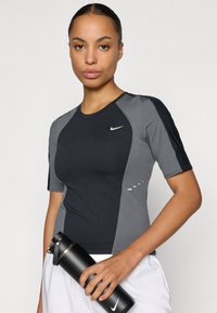 Αθλητικό τοπ σε μαύρο και γκρι με κοντά μανίκια, που διαθέτει λογότυπο Nike, καμπυλωτές ραφές και διακριτικές γραμμικές λεπτομέρειες. Πλαστικό μπουκάλι νερού κρατημένο στη μέση.
