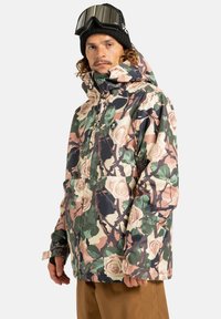Osoba ubrana w zimową kurtkę w kwiatowy wzór w odcieniach zieleni, beżu i różu, czarną czapkę beanie oraz gogle narciarskie na głowie.