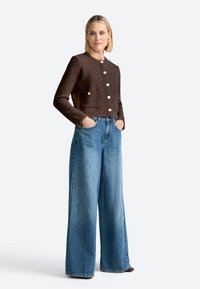 Giacca corta marrone con texture e bottoni dorati, dotata di due tasche frontali, abbinata a jeans in denim blu a gamba larga.