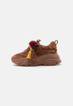 Baskets compensées en daim et tissu marron avec semelle épaisse, pompons décoratifs et glands sur les lacets, étiquetées Steve Madden sur le côté.