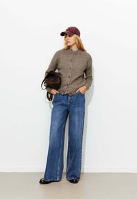 Cardigan en laine grise avec des boutons, associé à une chemise à carreaux, un jean en denim bleu large et une casquette bordeaux. Sac marron porté sur un bras.