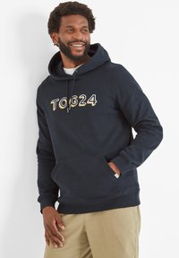 Smiling man with beard wearing navy hoodie with "TOG24" text, khaki pants, standing against white wall with one hand in pocket.