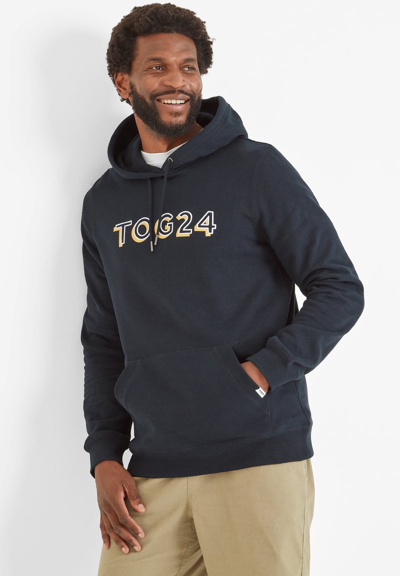 Smiling man with beard wearing navy hoodie with "TOG24" text, khaki pants, standing against white wall with one hand in pocket.
