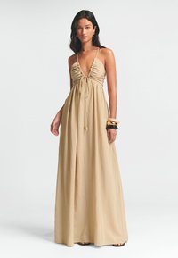Beige maxi-mekko, jossa on syvä V-pääntie, keräilty yksityiskohta rinnan kohdalla ja säädettävät olkaimet. Sujuva, kevyempi kangas, jossa on virtaava siluetti.