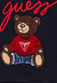 Design di un orso ricamato che indossa una maglietta rossa con un logo a triangolo e pantaloni blu, su uno sfondo scuro. Testo "indovina" in rosso sopra.