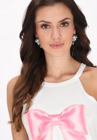 Fehér ujjatlan felső, amelyen egy nagy, rózsaszín selyemmasni grafika látható. A modellen túlnyúló gyöngy fülbevalók vannak, és hullámos, sötét barna haja.