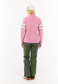 Pullover in pile rosa con strisce bianche sulle maniche, abbinato a pantaloni verdi. Il modello indossa un berretto crema e stivali beige con accenti marroni.
