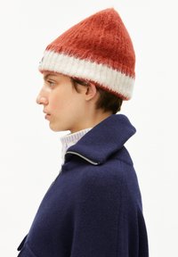 Persoon in profiel met een rood en wit gebreide muts, een blauwe ritssluitingjasje en een lichtgekleurde coltrui.