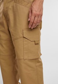 Hnědé cargo kalhoty s kapsou na stehně, s hladkou texturou a pevným šitím. Detailní záběr zdůrazňuje látku a designové detaily.