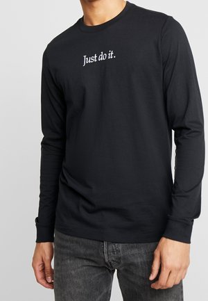 Personne portant un t-shirt noir à manches longues avec le texte blanc "Just do it.", associé à un jean noir délavé, debout devant un fond uni.