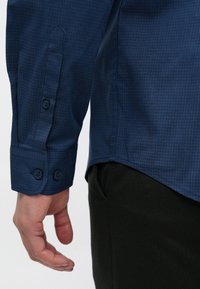 Sötétkék kockás ing gombos mandzsettával és alja lemetszett. Az anyag simának tűnik enyhe fényes felülettel.