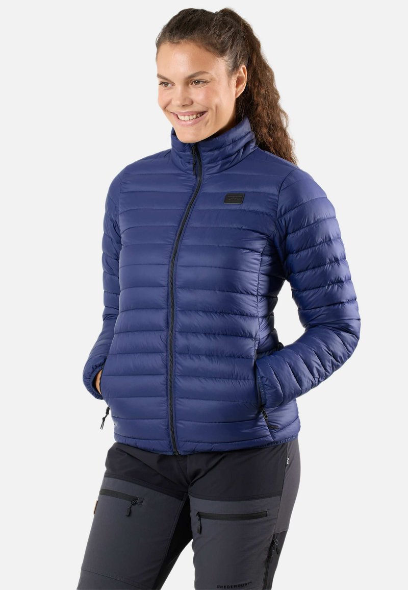 Tummansininen untuvapuku korkealla kauluksella, vetoketjulla ja vaakasuoralla tikkauskuviolla. Varustettu sivutaskuilla ja pienellä logopatchilla.