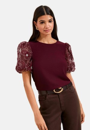 Kobieta w bordowej bluzce z przezroczystymi, haftowanymi kwiatowymi bufkami oraz ciemnych spodniach z czarnym paskiem z pozłacaną klamrą.